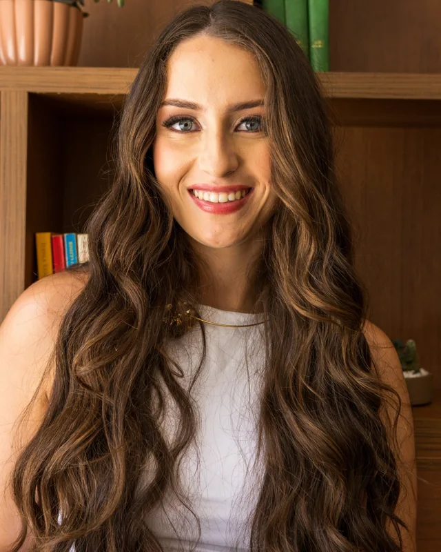 Retrato de Tarcylla Moura sorrindo, com o cabelo longo e ondulado, em frente a uma estante de madeira